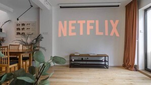 Ein Wohnzimmer mit einem Holzesstisch, Stühlen, einer TV-Konsole mit Büchern und einem an der Wand befestigten Projektor, der "NETFLIX" anzeigt.