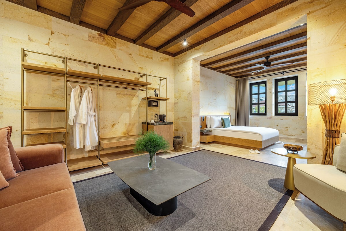 A modern hotel room with a large bed, a sitting area with a sofa, a coffee table, a wardrobe with hanging clothes, and a small table with a lamp.