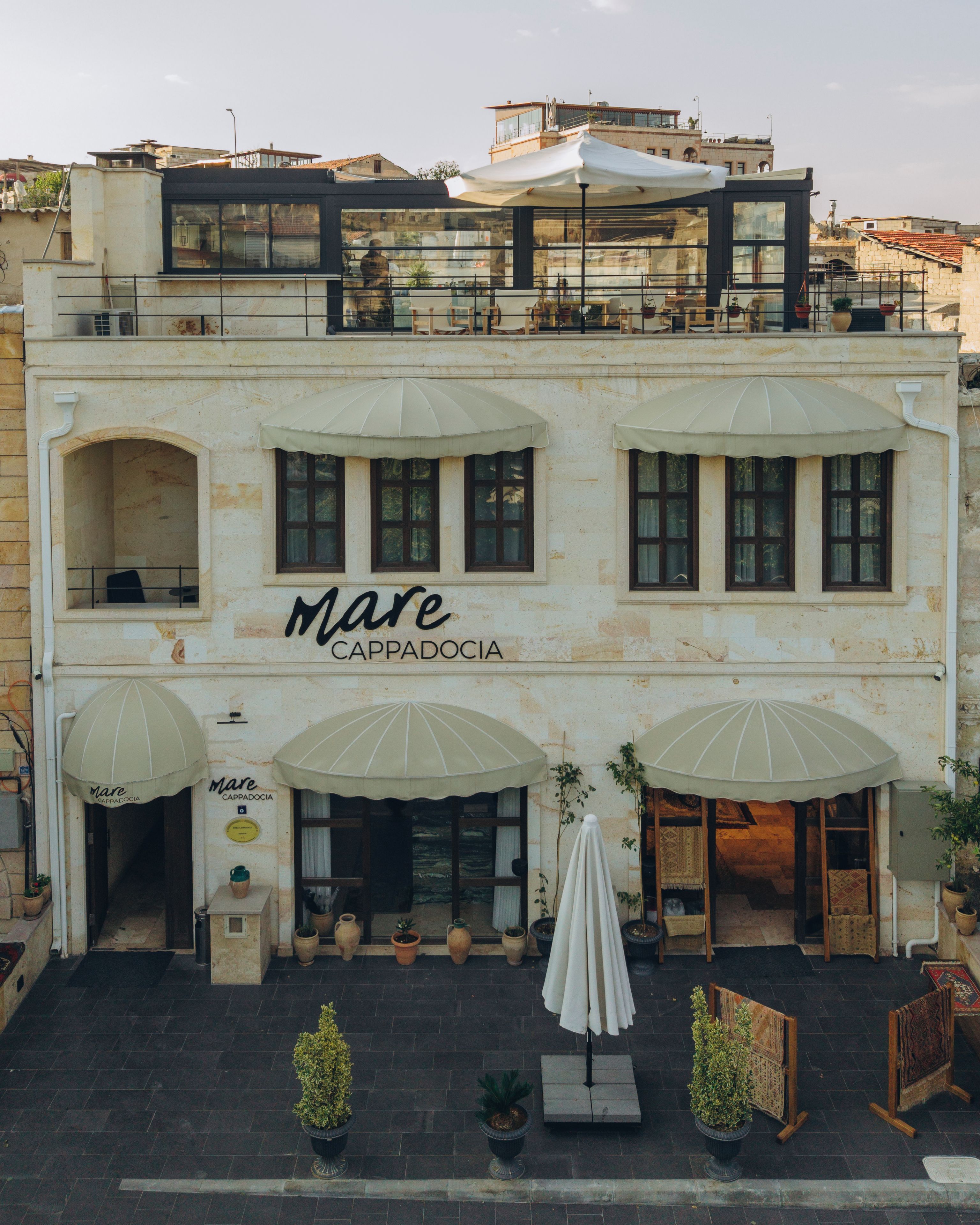 Un edificio con una cafetería llamada 'Mare Cappadocia' que cuenta con mesas al aire libre, sombrillas, plantas en macetas y fachada de piedra.
