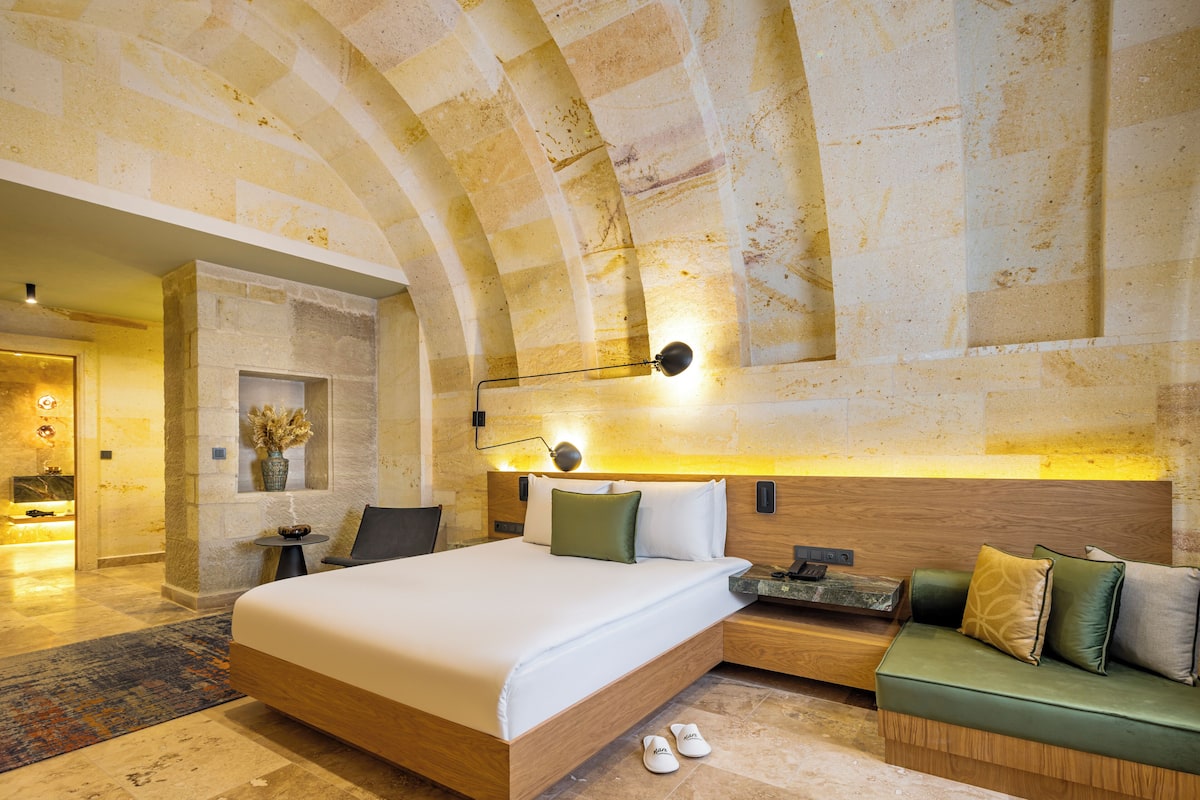 A modern hotel room with a large bed, a seating area, and a stone wall with a wooden headboard.