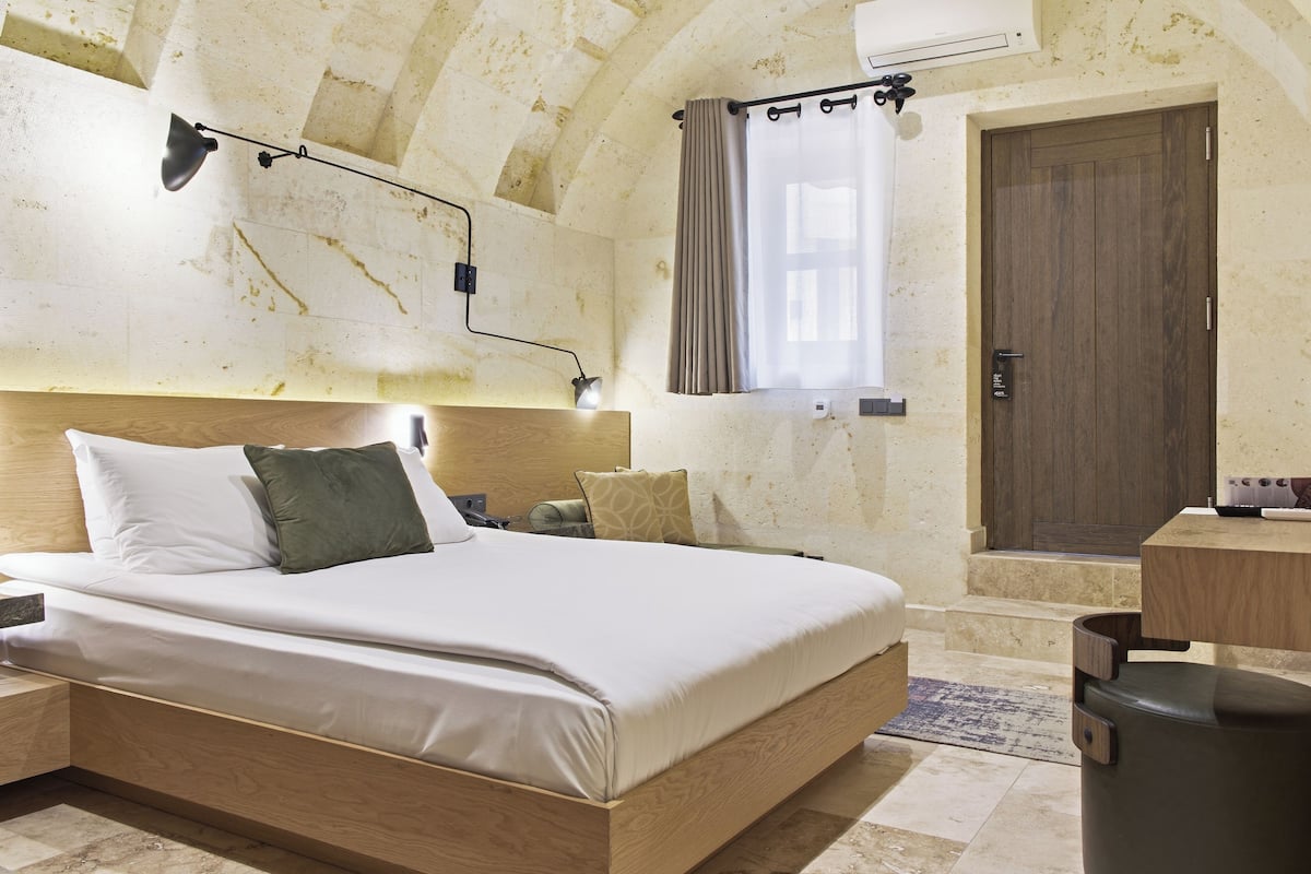 A bedroom with a wooden bed, a headboard, a nightstand, a wall-mounted lamp, a window with curtains, and a door.