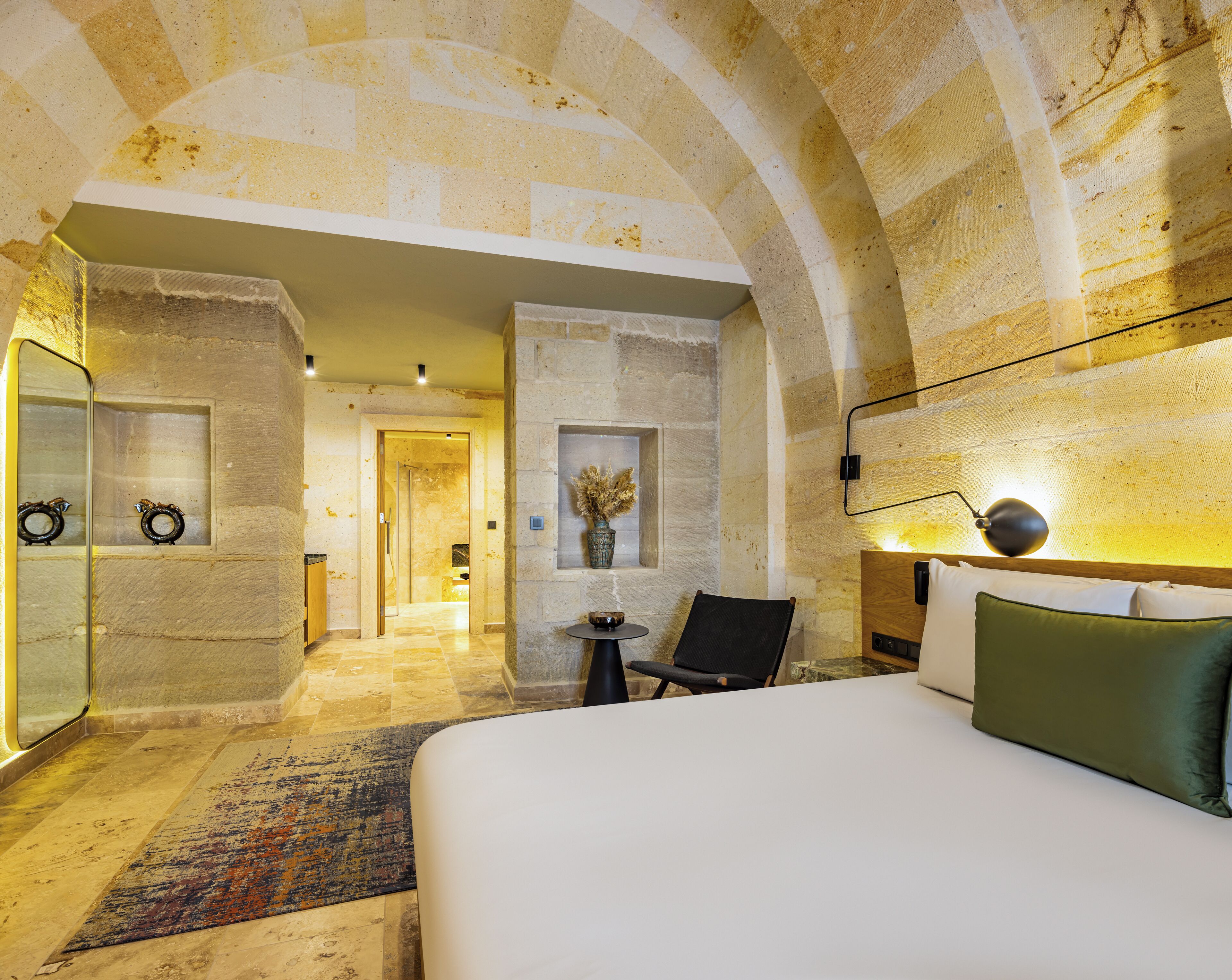 A hotel room with a large bed, a chair, a small table, and a mirror. There is a door leading to another room and a wall-mounted light fixture.