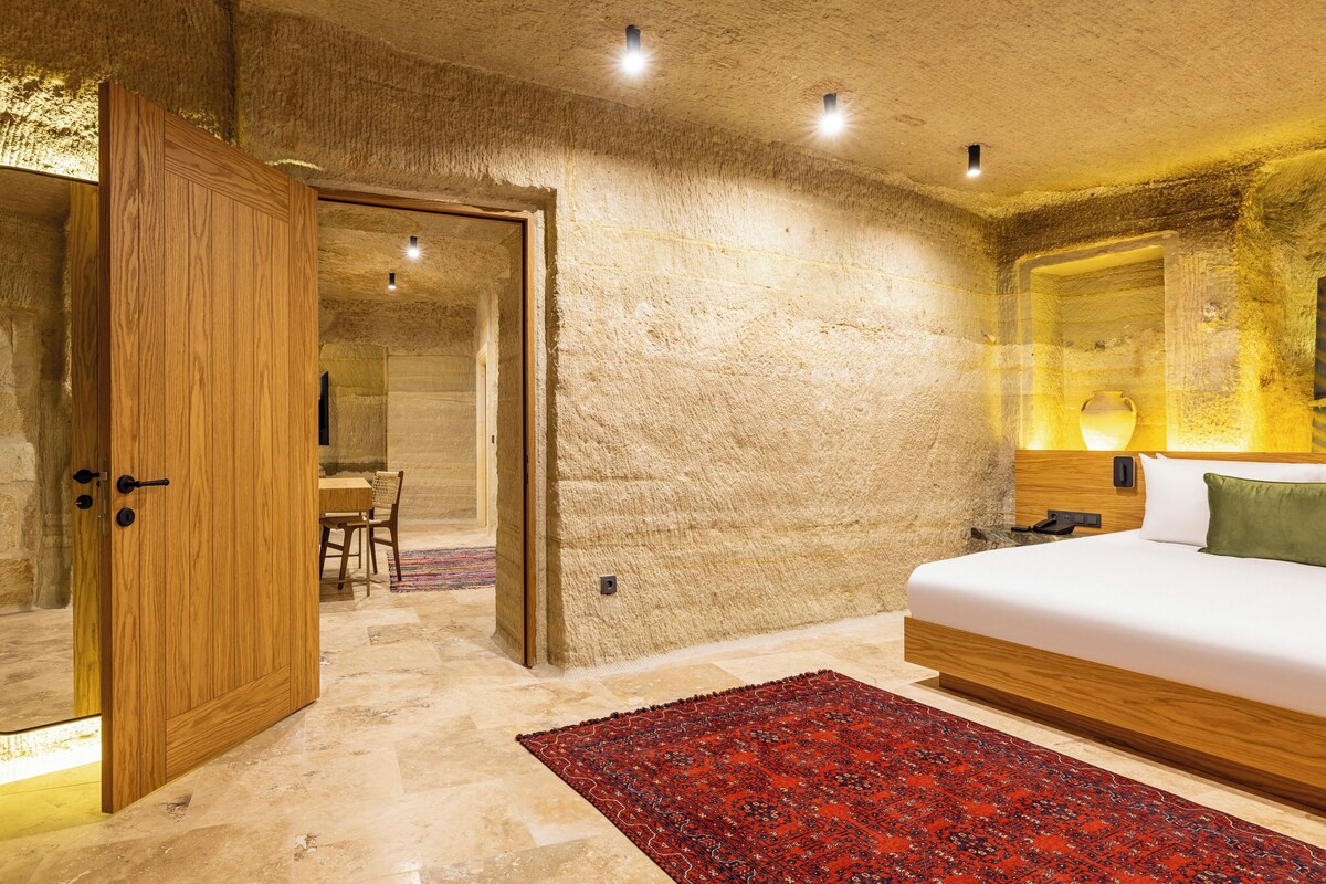 A bedroom with a wooden door, a bed with a white mattress, a red patterned rug, and a stone wall.