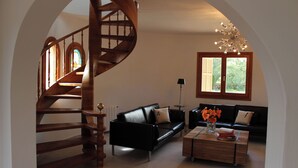 A spacious living area with a black leather sofa, a wooden coffee table with books and flowers, a black leather couch, a wooden spiral staircase, and a large window with a view of greenery. - Torre al Mar: sea view and pool, 150m to the beach, 8 pers., aircon, ETV/5162  (Cala Santanyí)