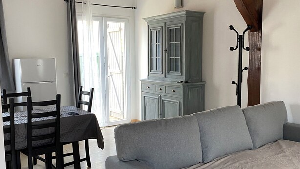A living room with a grey sofa, wooden beams, a dining table with chairs, and a white refrigerator. - Accommodation La Chaumière de Rambouillet 1-6 pers (Rambouillet)