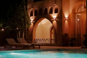 Ein Poolbereich mit Liegestühlen und einem Gebäude mit Rundbogenfenstern und warmer Beleuchtung.