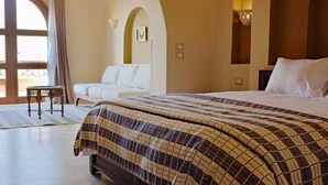 Habitación de hotel con una cama grande, un sofá y una mesita pequeña. Hay una ventana con cortinas, una lámpara de pared y un techo abovedado.