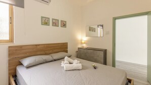 Ein ordentlich bezogenes Bett mit Holzkopfstück, ein Nachttisch mit Lampe und ein an der Wand befestigter Spiegel.