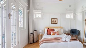 A bedroom with a large bed, two bedside tables, a chair, and a window with decorative glass. - Five Dock Family Home With Pool & Parking! (Five Dock)