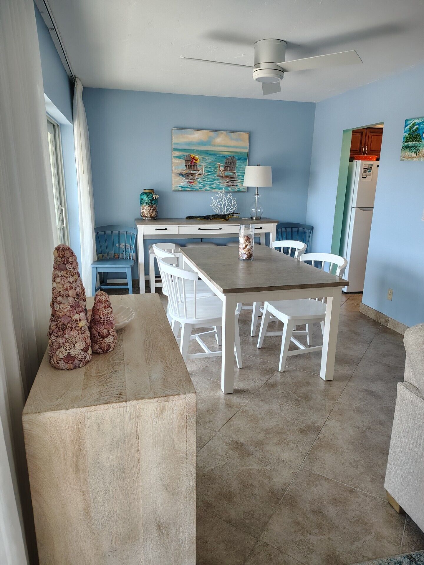 A dining area with a wooden table and white chairs, a ceiling fan, a painting of a beach scene, and a kitchen area with a refrigerator and cabinets.