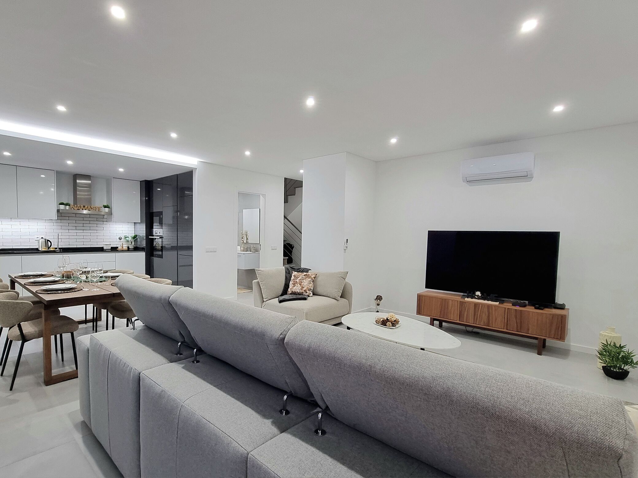A modern living room with a grey sectional sofa, a dining area with a table set for four, a flat-screen TV on a wooden stand, and a staircase leading to an upper level.
