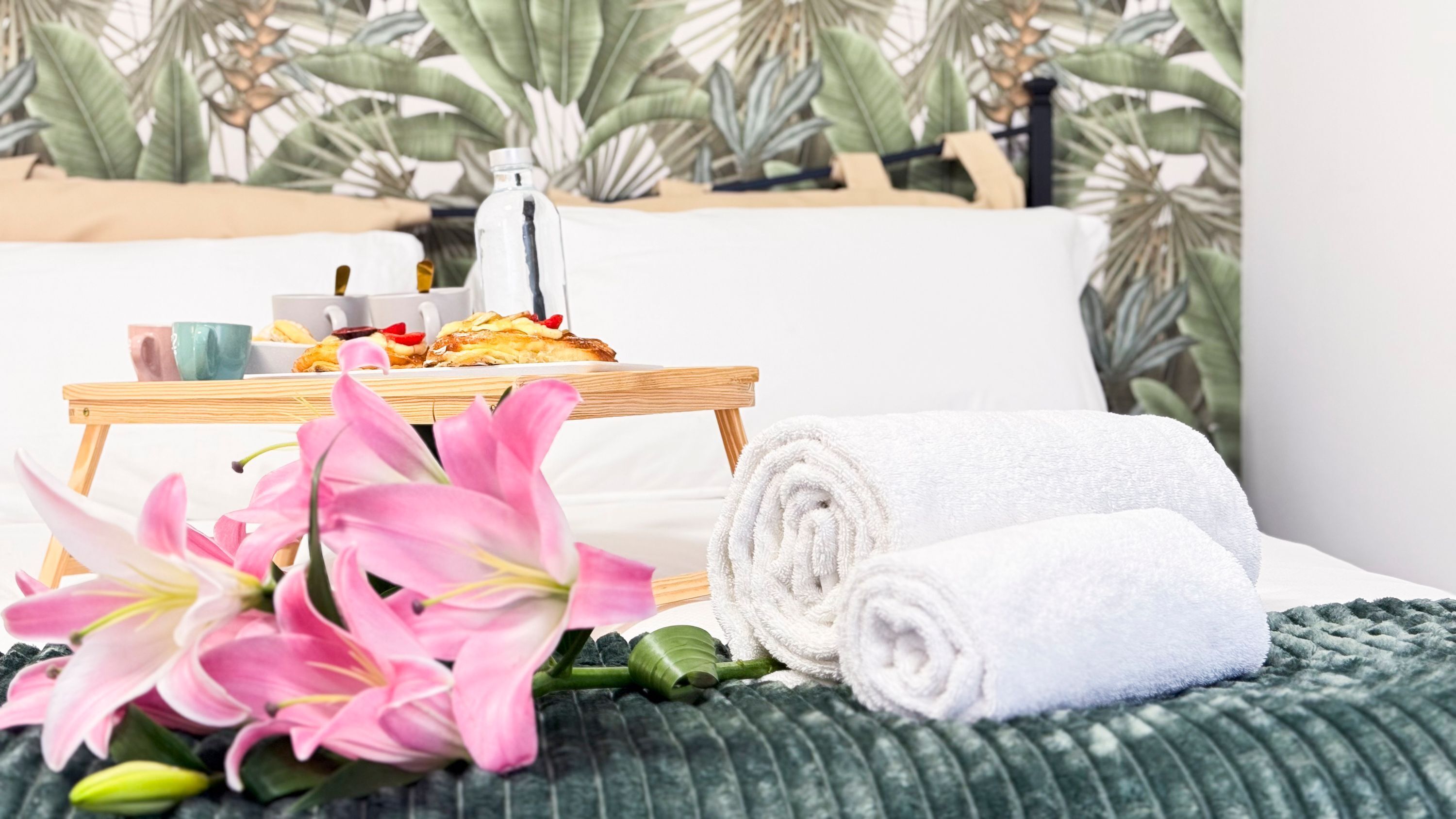 A neatly made bed with a green textured blanket, a wooden tray with pastries and a bottle, and pink flowers in the foreground.