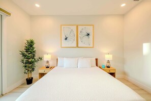 A bedroom with a large bed, two bedside tables with lamps, a potted plant, and two wall-mounted artworks.