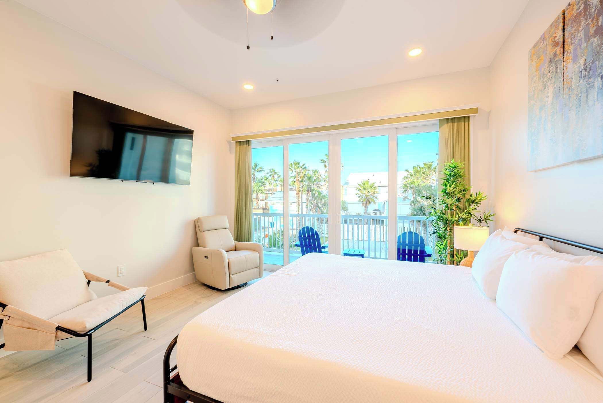 A hotel room with a large bed, a TV mounted on the wall, a seating area with a chair, and a balcony with a view of palm trees.