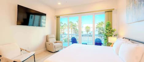 A hotel room with a large bed, a TV mounted on the wall, a seating area with a chair, and a balcony with a view of palm trees.