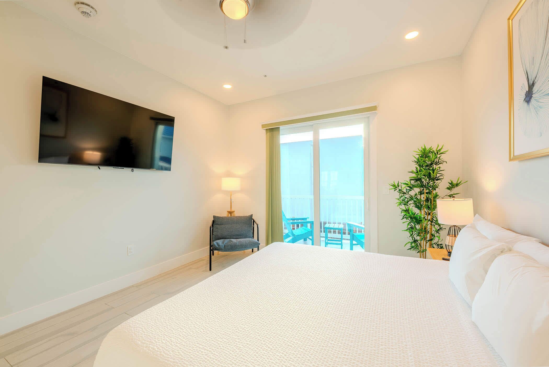 Une chambre d’hôtel avec un grand lit, un téléviseur à écran plat fixé au mur, une chaise, une plante en pot et un balcon avec des chaises bleues.