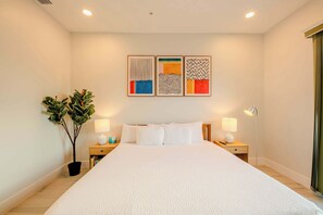 A modern hotel room with a large bed, two bedside tables with lamps, a potted plant, and three abstract wall art pieces.