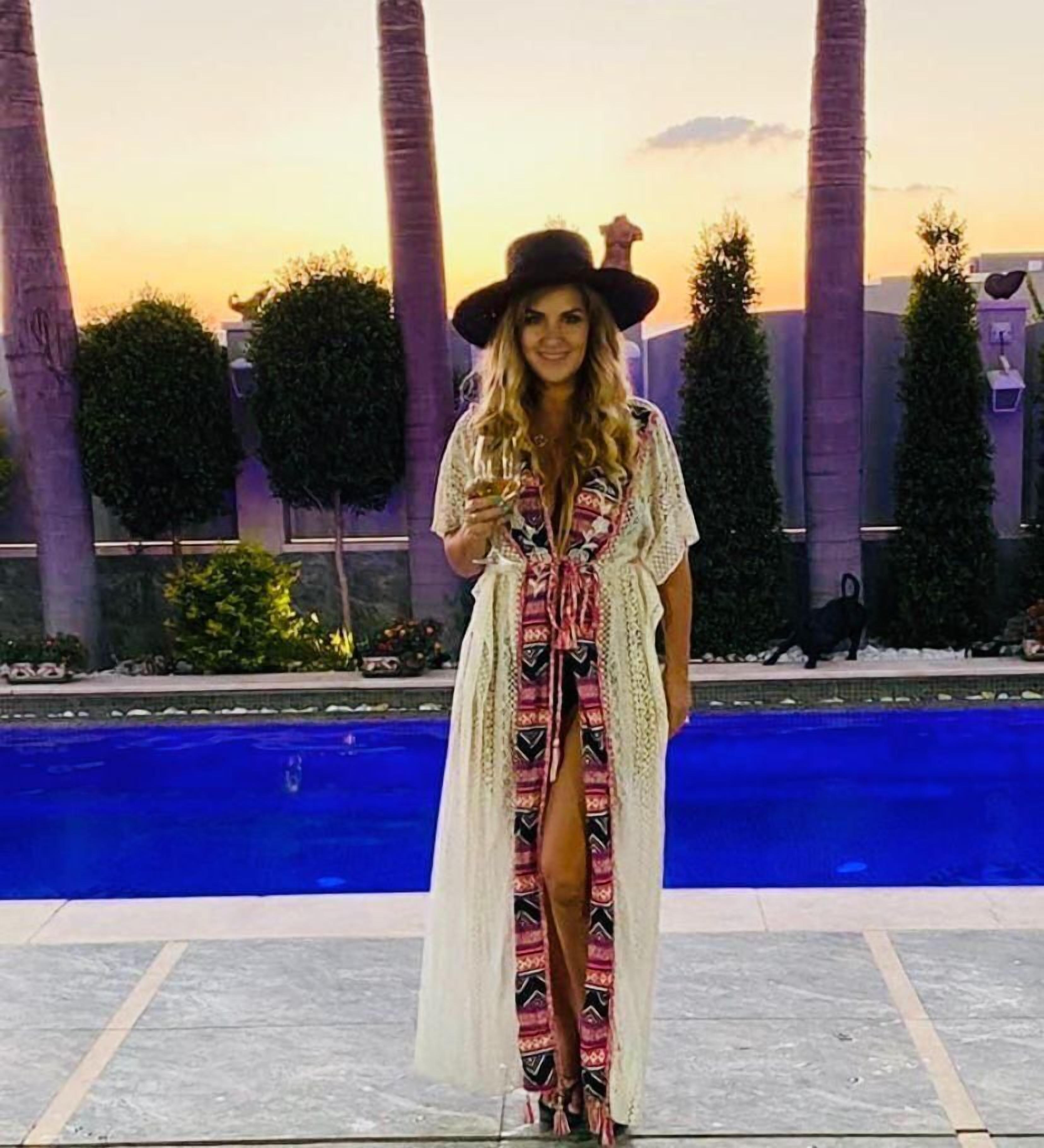 A woman standing by a pool with palm trees and a sunset in the background.