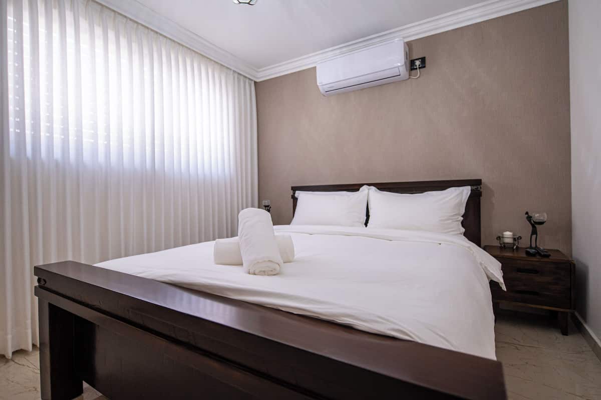 A neatly made bed with white linens and a rolled towel, a wooden nightstand with a lamp, and an air conditioning unit on the wall.
