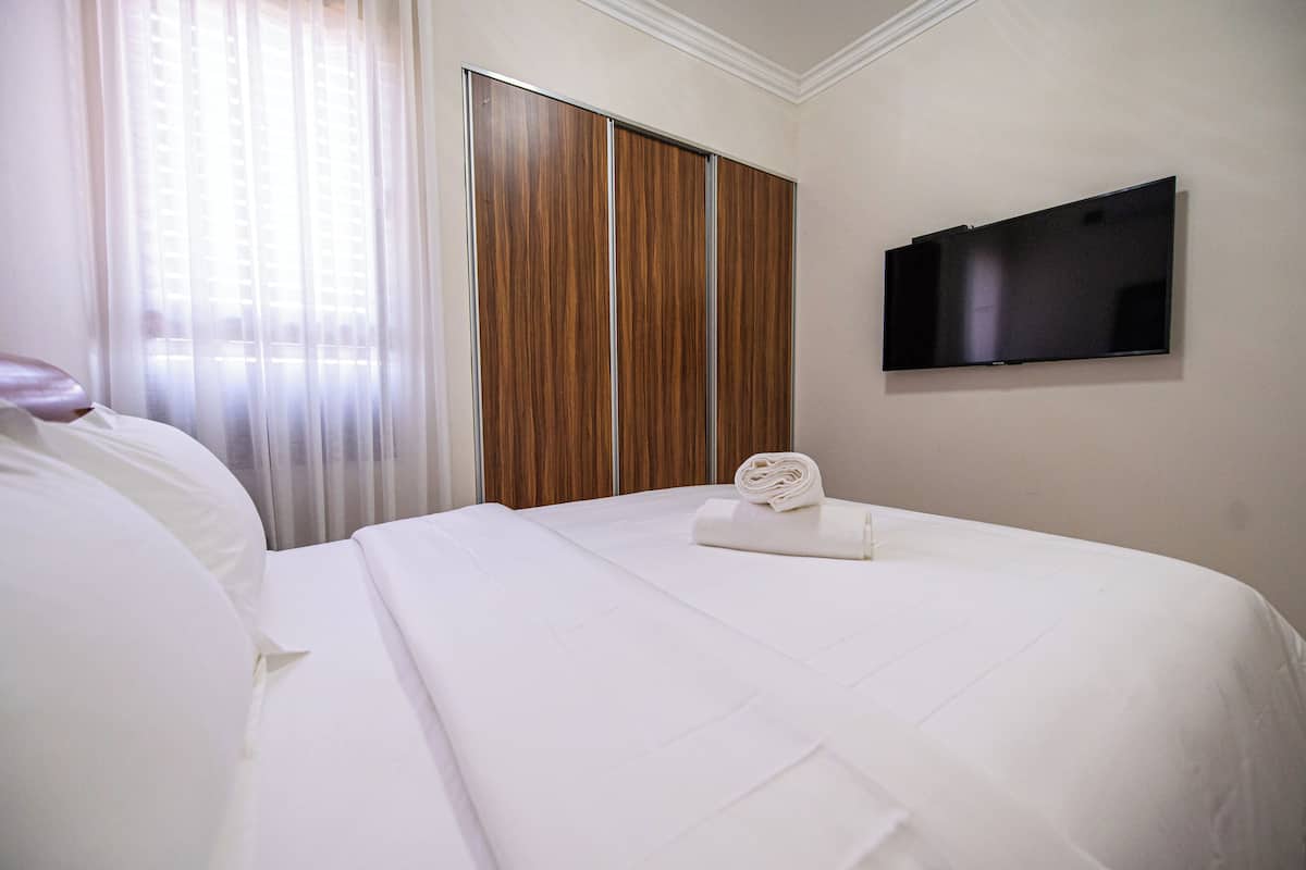 A hotel room with a bed, a TV mounted on the wall, and a window with sheer curtains.