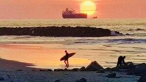 Een strandscène met een ondergaande zon, een schip op de achtergrond en iemand die een surfboard draagt.