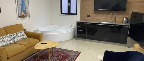 Una sala de estar con sofá, una mesita, un sillón azul, un ventanal grande, una cocina integrada con fregadero y un televisor fijado a la pared.