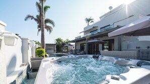 Een hottubgedeelte met een zwembad, lounge fauteuils en een modern gebouw met balkon.