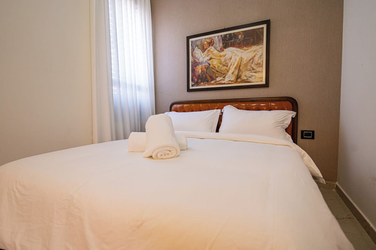 A neatly made bed with white linens and a rolled towel, a painting on the wall, and a window with curtains.