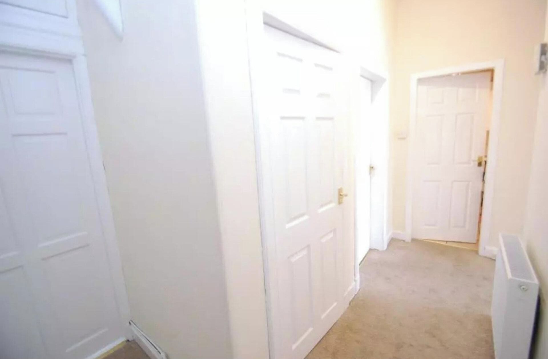 Un couloir avec des portes blanches, un sol recouvert de moquette et un radiateur sur le côté droit.