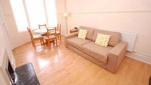 Un salon avec un canapé beige, deux coussins à motifs, une table à manger en bois avec des chaises et une petite table d’appoint.
