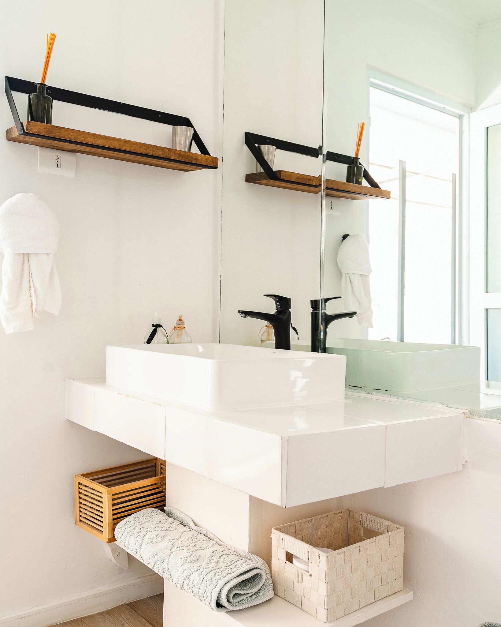Un baño con un lavamanos blanco, un grifo negro y un espejo encima. Hay repisas de madera sobre el lavamanos y una cesta tejida debajo.