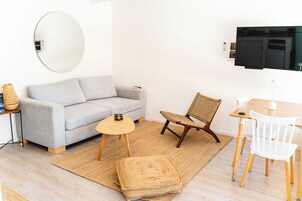 Una sala de estar moderna con un sofá gris, un espejo redondo, un televisor y una pequeña mesa de madera con sillas.
