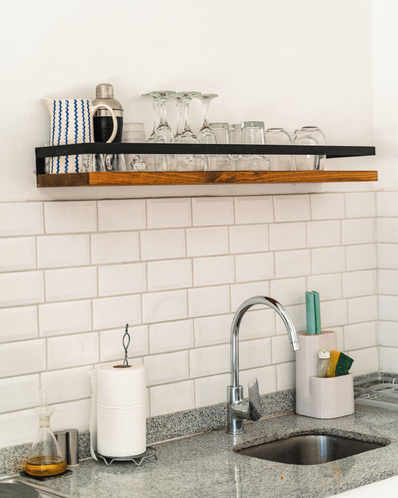 A kitchen with a granite countertop, a stainless steel sink, a faucet, a shelf with glassware, and a roll of paper towels.