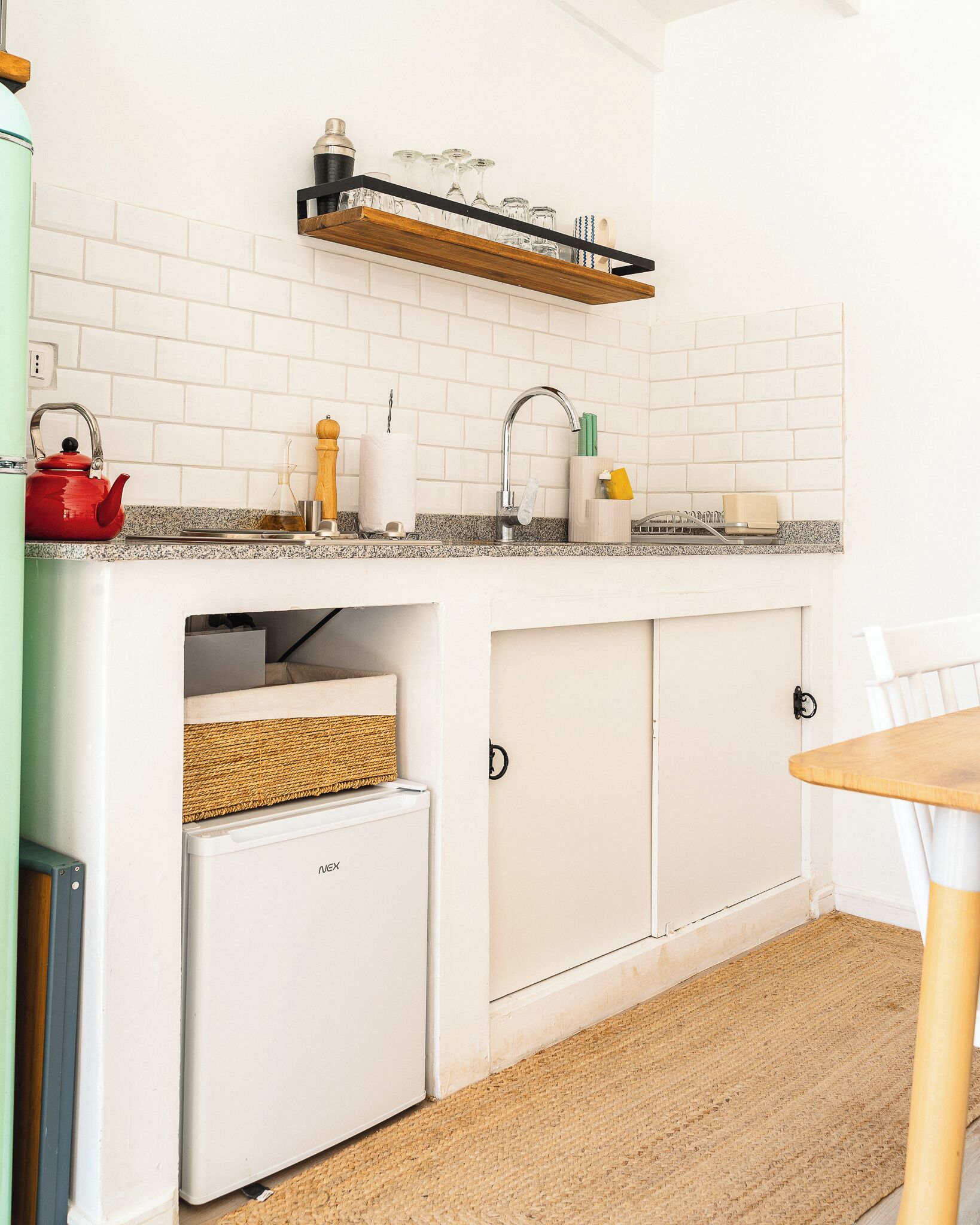 Een kleine keuken met witte kasten, een inbouwkoelkast, een spoelbak met een houten plank erboven en een tafeltje met een stoel.