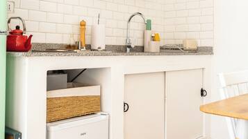 Een kleine keuken met witte kasten, een inbouwkoelkast, een spoelbak met een houten plank erboven en een tafeltje met een stoel.
