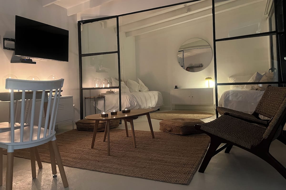 A modern bedroom with a large window, a flat-screen TV, a wooden table, and a comfortable chair.