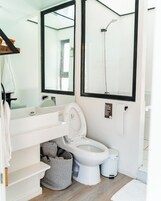 Un baño moderno con lavamanos, inodoro y ducha blancos. Hay un perchero y un cesto de basura.