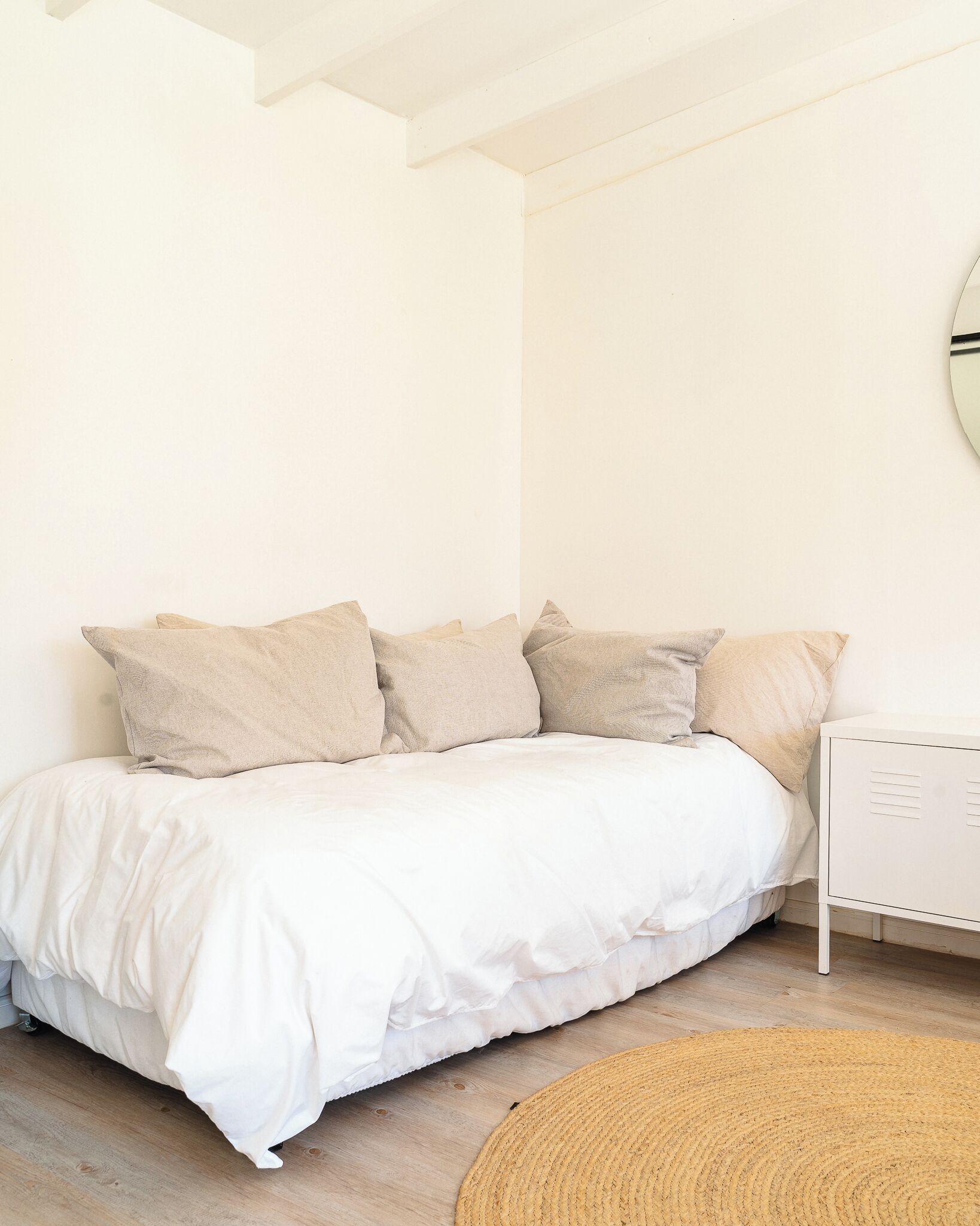 Una cama con ropa de cama blanca y varios almohadones color beige, un tapete redondo tejido y una mesita de noche blanca con un espejo.