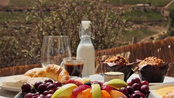 Un desayuno con cruasanes, frutas y bebidas sobre una mesa con vista a un viñedo.