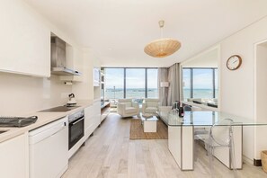 A modern kitchen with a view of the sea, a dining area with a glass table, and a living space with a sofa and chairs.