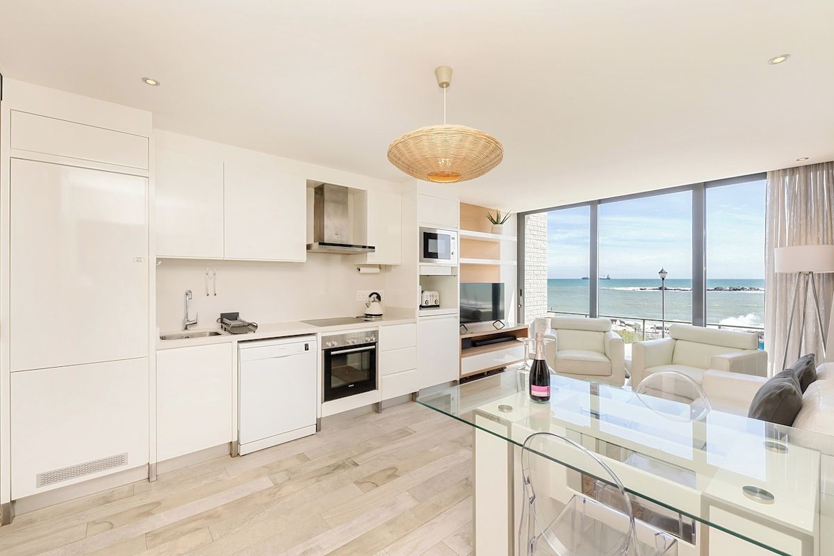 A modern kitchen with white cabinetry, a glass dining area, and a view of the sea.