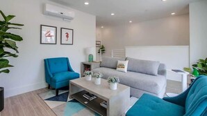 Una sala de estar moderna con un sofá gris, un sillón azul y una mesa de centro. Hay dos cuadros en la pared, una planta en maceta y una pequeña mesita auxiliar.