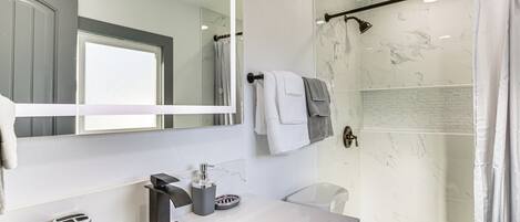 Un baño moderno con un espejo grande, un lavabo con encimera de mármol, ducha con puerta de vidrio y un perchero con varias toallas.