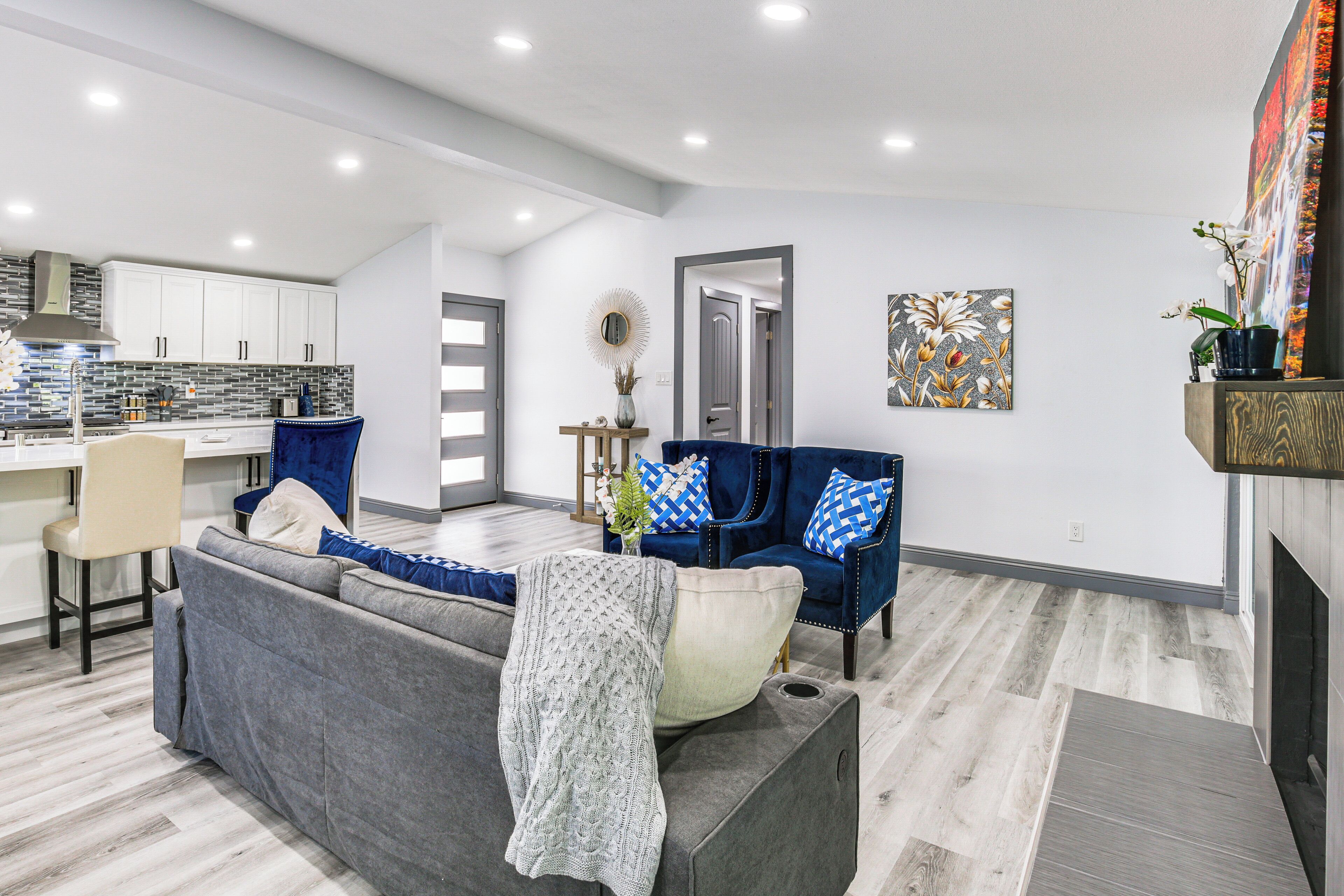 Una sala de estar moderna con un sofá gris, cojines azules y blancos, y una mesa de centro. Se ve una cocina al fondo con armarios blancos y un salpicadero de azulejos.