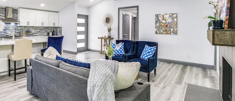 Una sala de estar moderna con un sofá gris, cojines azules y blancos, y una mesa de centro. Se ve una cocina al fondo con armarios blancos y un salpicadero de azulejos.