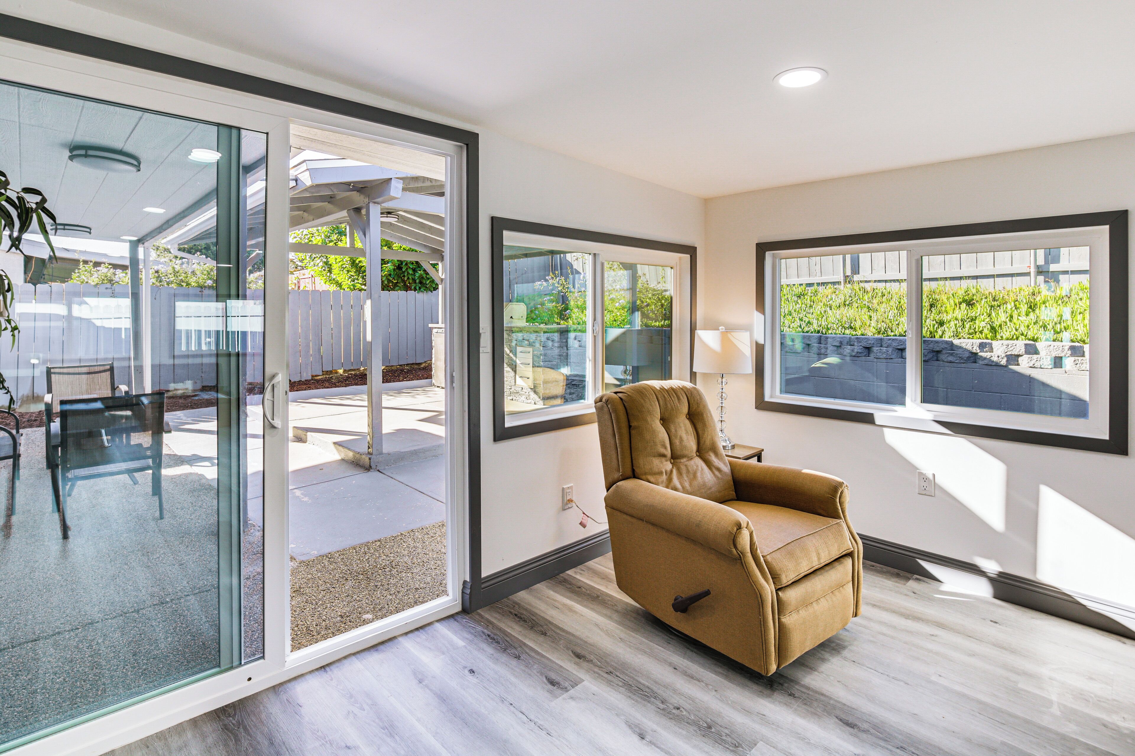 A modern living room with a sliding glass door leading to an outdoor patio, a leather armchair, and large windows offering views of greenery.