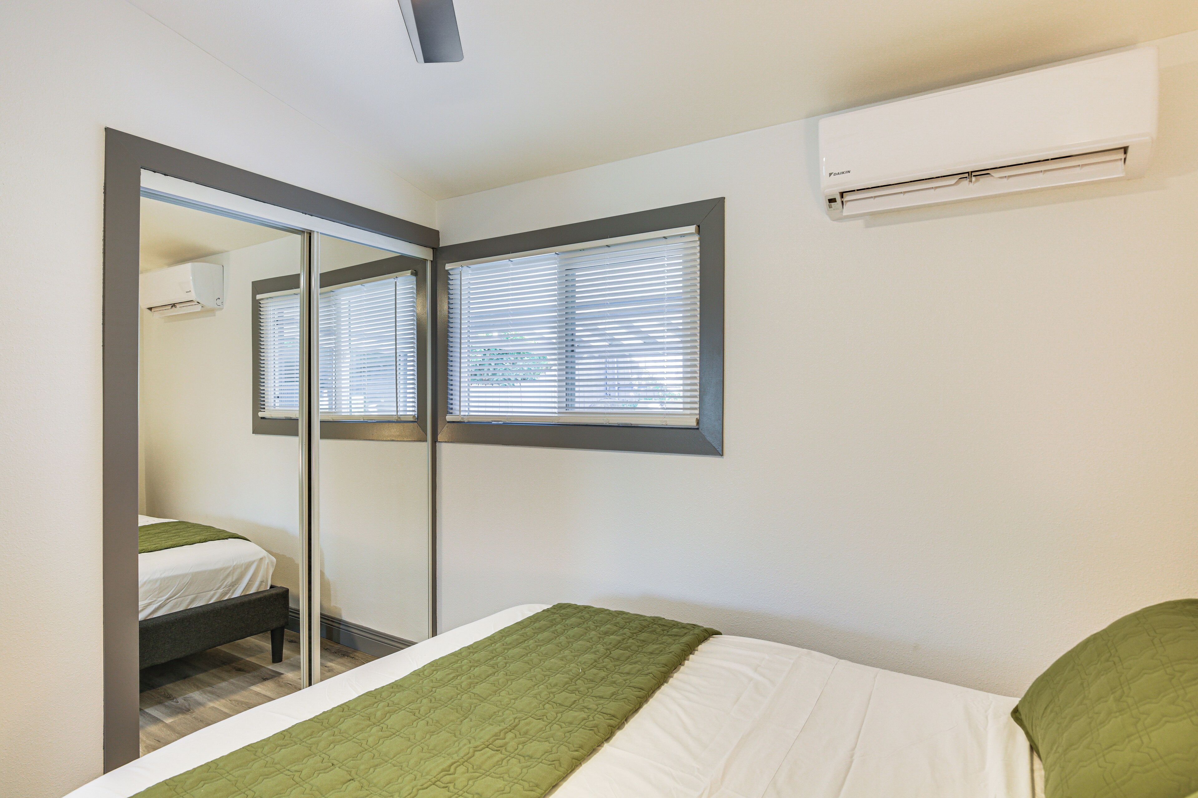 Un dormitorio con cama, espejo, ventana con persianas y un equipo de aire acondicionado en la pared.