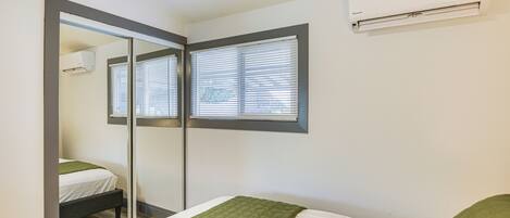 Un dormitorio con cama, espejo, ventana con persianas y un equipo de aire acondicionado en la pared.