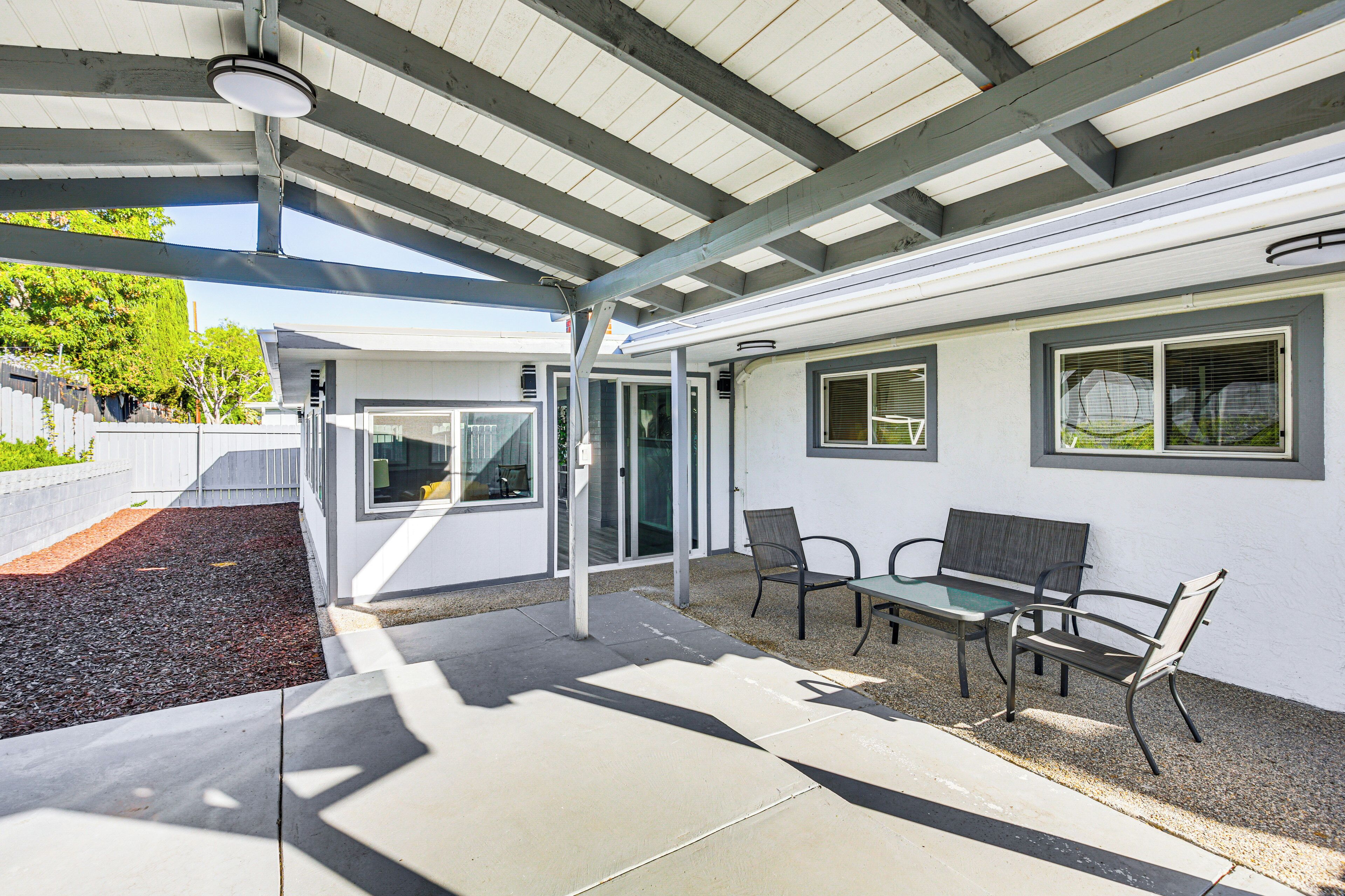 An outdoor patio area with a covered seating area, a concrete floor, and a gravel path leading to a house entrance.