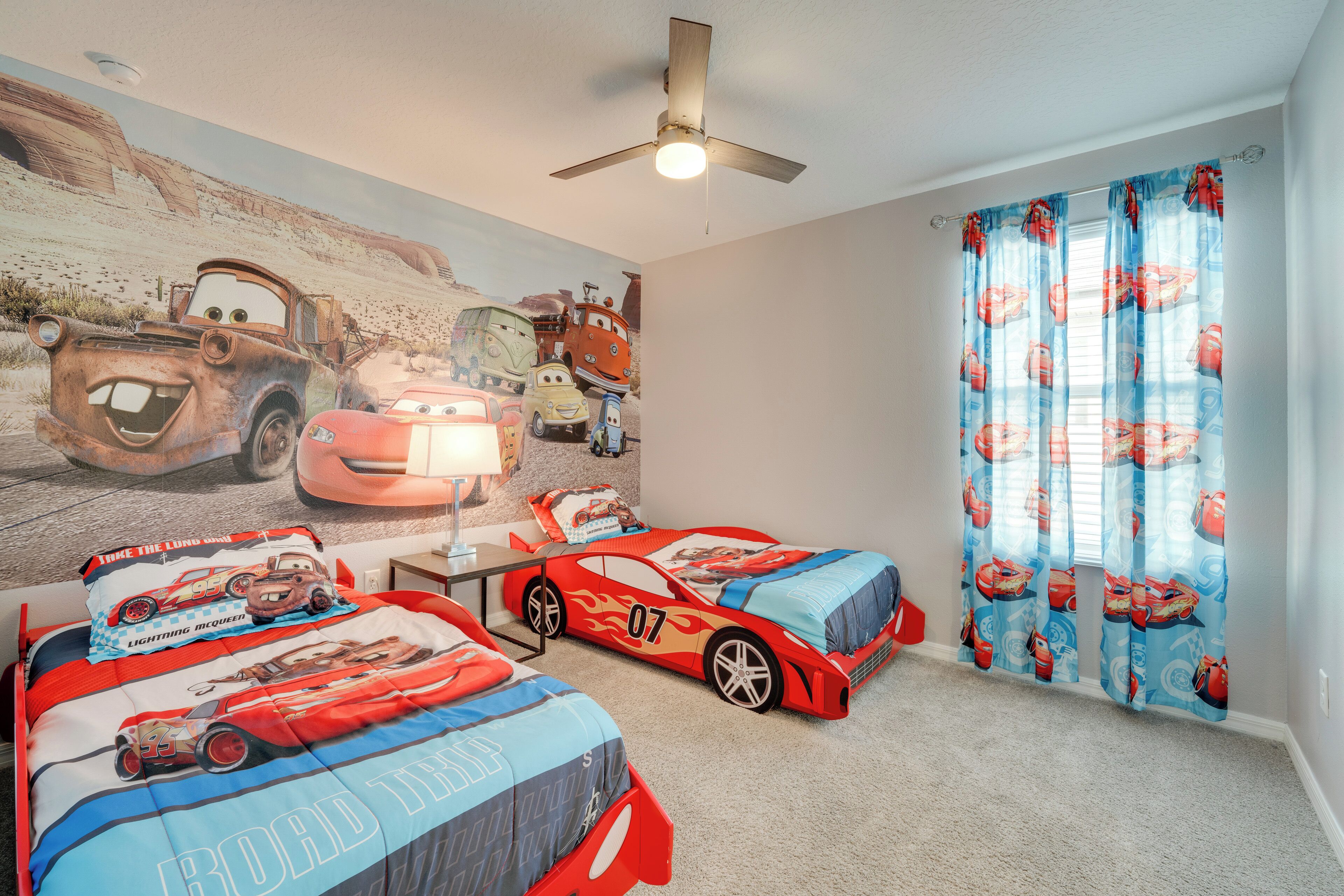 Een slaapkamer met twee bedden, een plafondventilator, een muurschildering van auto's en een raam met gordijnen.